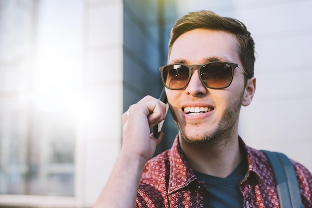 Primer plano retrato de feliz alegre atractivo hombre caucásico con elegantes gafas de sol en la ciudad Joven hombre de negocios sonríe y habla con un teléfono inteligente en la construcción al aire libre Concepto de estilo de vida urbano de moda