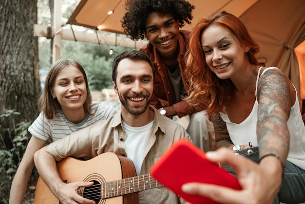 Primer plano retrato de un grupo de jóvenes amigos viajando juntos en glamping en el bosque divirtiéndose tocando la guitarra, haciendo selfie, transmitiendo a las redes sociales sentados en la carpa riendo