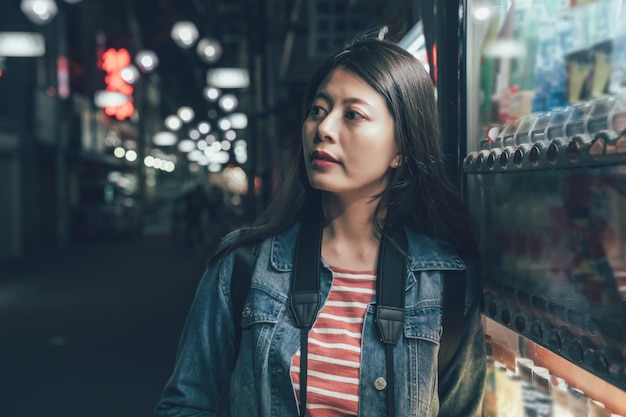 primer plano retrato de una hermosa mujer asiática viajera relajándose apoyándose en la máquina expendedora osaka Japón. joven turista con cámara mirando a un lado de pie en una calle tranquila por la noche en la ciudad.