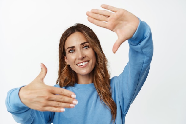 Primer plano retrato de una hermosa mujer de mediana edad mira a través de los marcos de la mano y sonríe, imaginando algo, buscando el ángulo perfecto, fondo blanco.