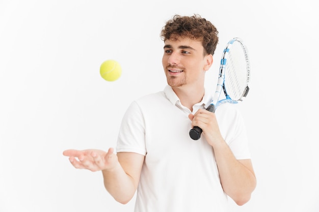 Primer plano retrato de hombre alegre en camiseta sosteniendo la raqueta y la pelota mientras juega al tenis aislado sobre la pared blanca