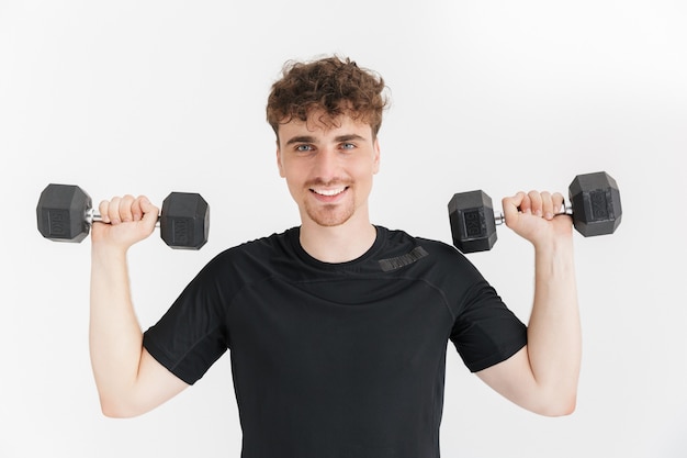 Primer plano retrato de hombre deportivo satisfecho en camiseta mirando al frente y levantando pesas durante el entrenamiento aislado sobre la pared blanca