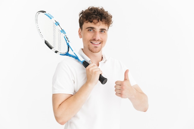 Primer plano retrato de hombre feliz en camiseta mostrando el pulgar hacia arriba y sosteniendo la raqueta mientras juega tenis aislado sobre la pared blanca