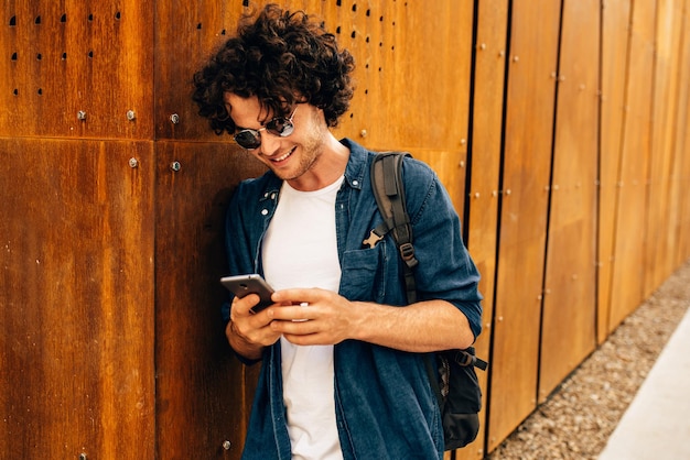 Primer plano retrato de hombre guapo de pie al aire libre escribiendo mensajes en el teléfono móvil Hombre joven con cabello rizado usa gafas de sol descansando afuera en la ciudad navegando en su teléfono celular en la pared moderna