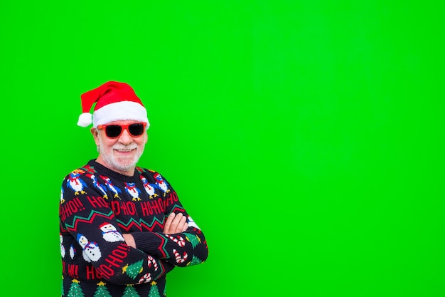 Primer plano y retrato de hombre maduro y anciano o senior sonriendo y mirando a la cámara vistiendo ropa de navidad y sombrero con fondo verde con espacio de copia