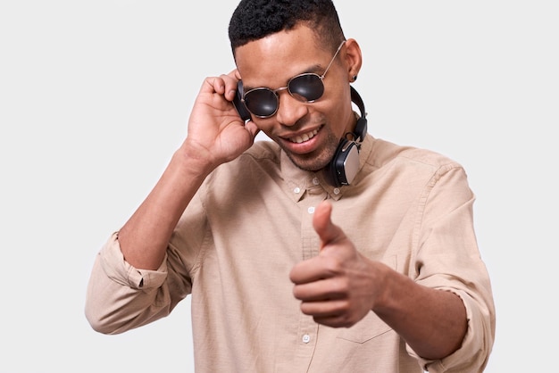 Primer plano retrato de un joven afroamericano con auriculares escuchando la música y bailando Feliz sonriente DJ afro masculino con gafas de sol de espejo y ropa informal posando sobre la pared blanca del estudio