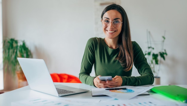 Primer plano retrato de una joven y encantadora mujer de negocios confiada que usa un teléfono inteligente mientras trabaja con una computadora portátil en casa