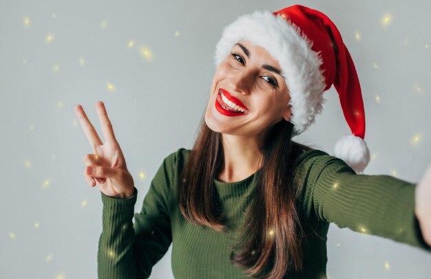 Primer plano retrato de una joven muy linda con gesto de paz con sombrero navideño de Papá Noel y tomándose un selfie aislado sobre fondo gris Feliz año nuevo 2024 y feliz Navidad