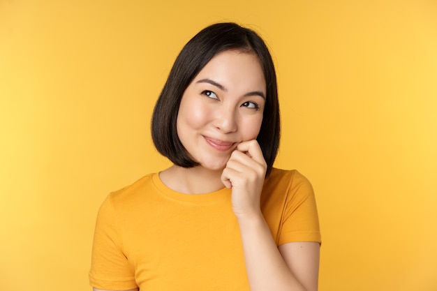 Primer plano retrato de linda chica asiática sonriendo pensando mirando hacia arriba pensativo de pie en camiseta sobre fondo amarillo Copiar espacio