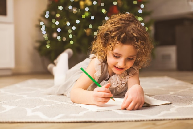 Primer plano retrato de linda niña rizada acostada en un piso frente al árbol de Navidad Niña preescolar escribiendo su carta de deseo a Santa en la víspera de Navidad