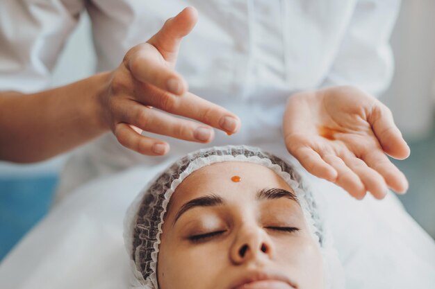 Primer plano retrato de manos de esteticista goteando aceite nutritivo en la frente y la piel facial de jóvenes...