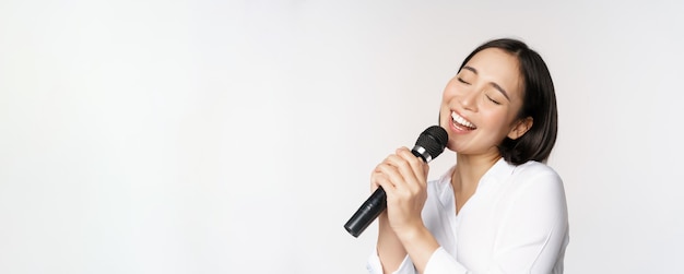 Primer plano retrato de mujer asiática cantando en el micrófono en el karaoke de pie sobre fondo blanco.