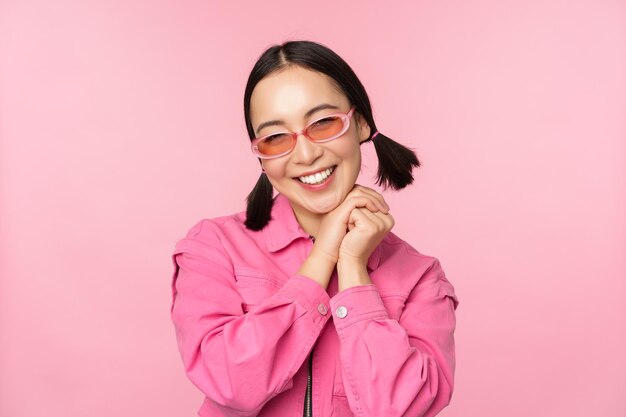 Primer plano retrato de mujer asiática con estilo en gafas de sol sonriendo mirando halagado riendo coqueto de pie sobre fondo rosa Copiar espacio