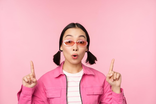 Primer plano retrato de una mujer asiática jadeando sorprendida señalando con el dedo el anuncio de pancarta de pie sobre fondo rosa