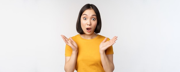 Primer plano retrato de una mujer asiática que parece sorprendida cara de asombro mirando impresionada a la cámara de pie sobre fondo blanco en camiseta amarilla