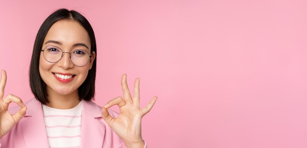 Primer plano retrato de una mujer corporativa impresionada mujer de negocios asiática con gafas que muestra un signo de estar bien mirando asombrado a la cámara recomendando fondo rosa