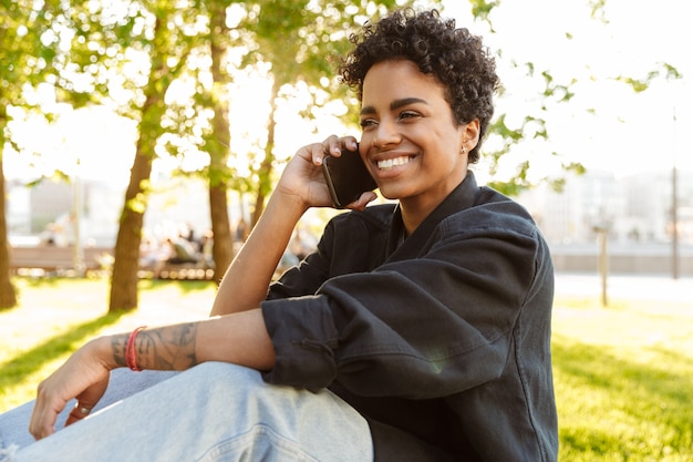Foto primer plano retrato de mujer feliz con pelo rizado hablando por teléfono inteligente mientras está sentado en un banco en el parque de la ciudad