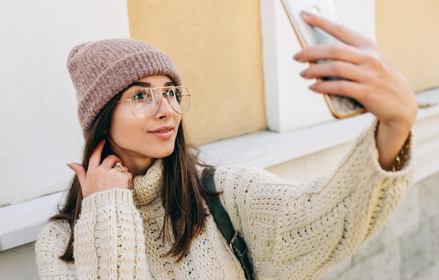 Primer plano retrato de mujer joven de pie al aire libre contra la pared del edificio y tomando autorretrato en el teléfono inteligente Hermosa mujer vestida con anteojos de sombrero de suéter de punto haciendo selfie