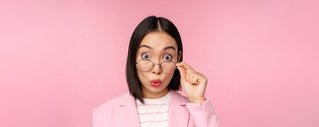 Primer plano retrato de la señora de la oficina coreana se quita las gafas y mira impresionado a la cámara expresión de la cara sorprendida fondo rosa