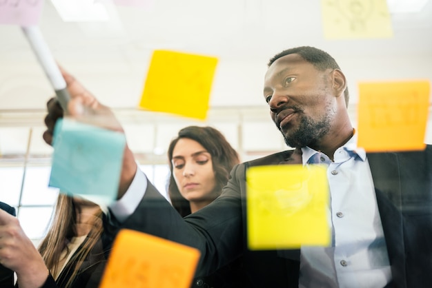 Primer plano de una reunión de lluvia de ideas de empresario africano sonriente con sus colegas mediante el uso de una colorida nota de papel adhesivo en la pared de vidrio para encontrar nuevas ideas.