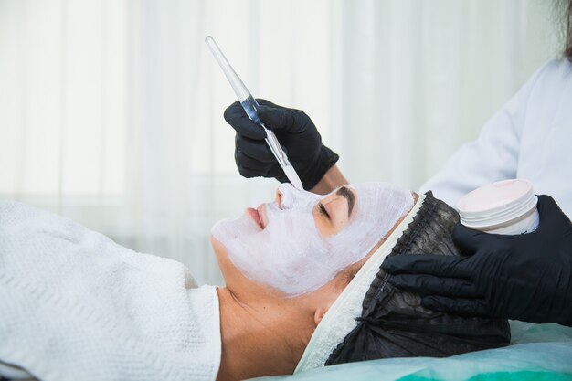 Primer plano de rostro de mujer en procedimiento de máscara de arcilla blanca en salón de belleza. Mascarilla exfoliante facial, tratamiento de belleza spa, concepto de cuidado de la piel.