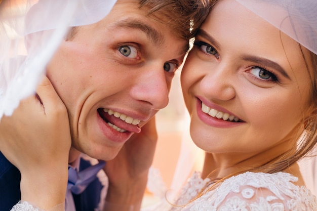 Foto primer plano de los rostros de los recién casados bajo el velo de novia, el novio muestra la lengua y la sonrisa de la novia