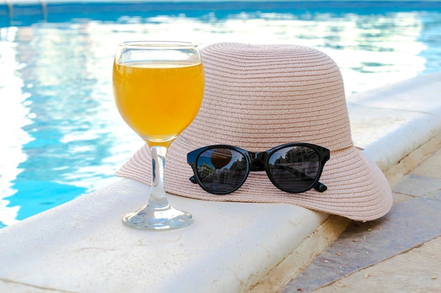 Primer plano de sombrero de paja amarillo y gafas de sol protectoras negras gafas de jugo de naranja fresco batido bebida cóctel en la piscina del hotel con agua azul en un día cálido y soleado Concepto de vacaciones de verano