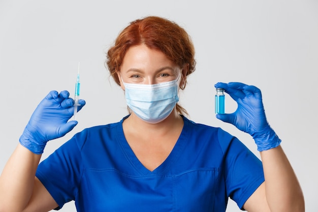 Primer plano de sonriente doctora amable en mascarilla y guantes de goma con vacuna de virus y jeringa, haciendo tiro, inyección.