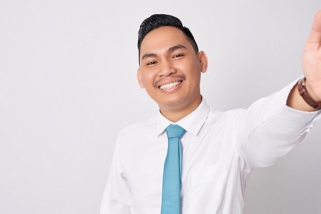 En primer plano sonriente, feliz, divertido, joven empresario asiático con una camisa formal y corbata aislado sobre un fondo blanco tomando una selfie en el teléfono