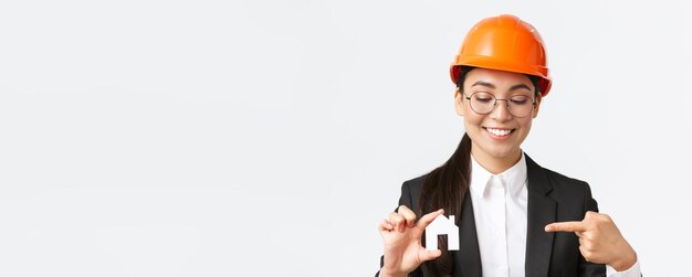 Primer plano de una sonriente ingeniera arquitecta asiática con casco y traje de negocios sosteniendo una miniatura casera y señalando con el dedo a una pequeña casa trabajando sobre la construcción de edificios de fondo blanco