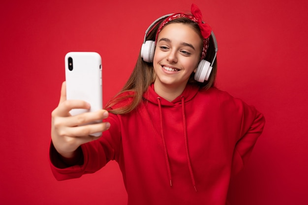 Primer plano de una sonriente niña bonita morena con sudadera con capucha roja aislada sobre fondo rojo sosteniendo