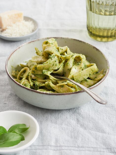 Foto primer plano de un tazón de pasta verde con un tenedor sobre pesto de tagliatelle verde junto a hojas de albahaca y queso parmesano comida tradicional italiana