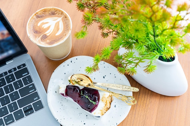 Primer plano del teclado de la computadora portátil Café con leche caliente con espuma de leche de arte latte en una taza hecha de papel Pastel de arándanos en el escritorio de madera en la vista superior Como desayuno En una cafetería durante el concepto de trabajo empresarial