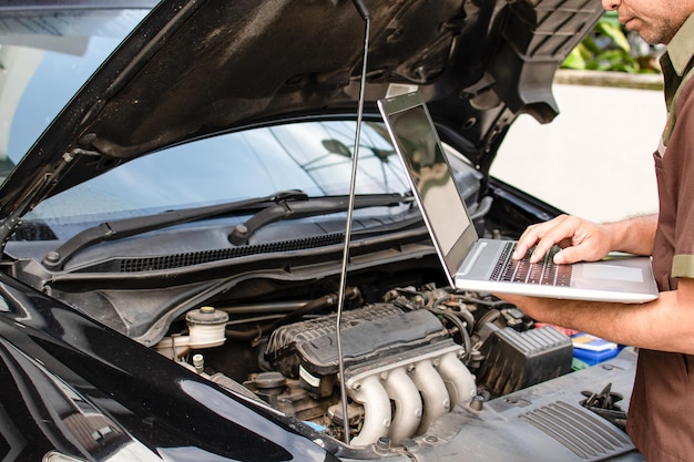 Primer plano Un técnico masculino sostiene una computadora portátil para comprobar el funcionamiento del motor de los automóviles negros en el garaje El motor de un automóvil no se enciende Hombre usando un ordenador portátil Fondo borroso