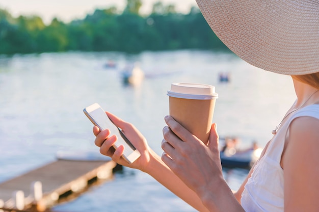 Primer plano, un teléfono inteligente con una taza de café en manos de una niña. Al fondo hay un terraplén con agua y yates, botes, catamaranes.