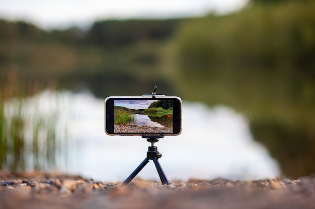 Un primer plano del teléfono en un trípode toma un video o una foto de la naturaleza. Un hermoso lago en el bosque con nubes en la pantalla del teléfono móvil del fotógrafo.