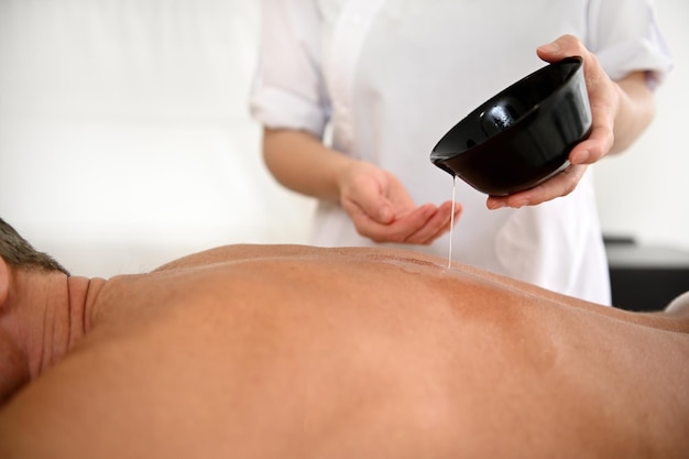 Primer plano de un terapeuta de masaje vertiendo un poco de aceite del recipiente en la espalda de un hombre maduro acostado en la mesa de masajes durante una sesión de terapia de masaje en la clínica de spa de salud. Concepto de bienestar y cuidado corporal.