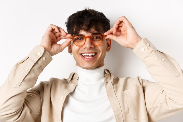 Primer plano de tipo hipster con estilo probando gafas en la tienda de óptica, ponerse gafas y sonriendo, de pie en la pared blanca.