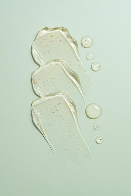 Primer plano transparente de gotas de suero de oro de 24k en fondo verde que muestra la textura de gel de crema cosmética clara exprimida con efectos de brillo sutiles