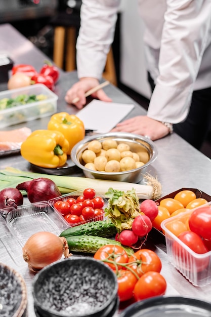 Primer plano de verduras frescas en la mesa en la cocina comercial con el chef haciendo una lista en segundo plano.