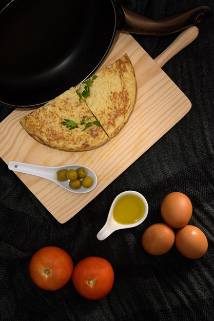 Foto un primer plano vertical de alto ángulo de dos piezas de tortilla en una mesa de cocina de madera con aceitunas, yemas de huevo, tomates y huevos frescos en los lados