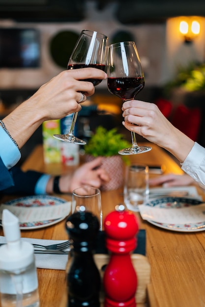 Primer plano vertical de pareja irreconocible tintineo de vasos con vino tinto sentado en la mesa en el restaurante por la noche