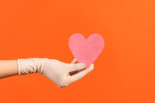 Primer plano de la vista lateral de la mano humana en guantes quirúrgicos blancos sosteniendo en la mano una pequeña forma de corazón rosa.