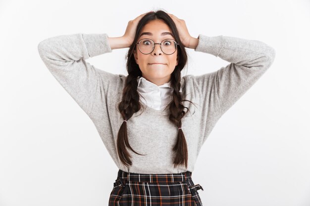 Primer retrato de una adolescente asombrada con anteojos y uniforme escolar preguntándose mientras agarra su cabeza aislada sobre la pared blanca