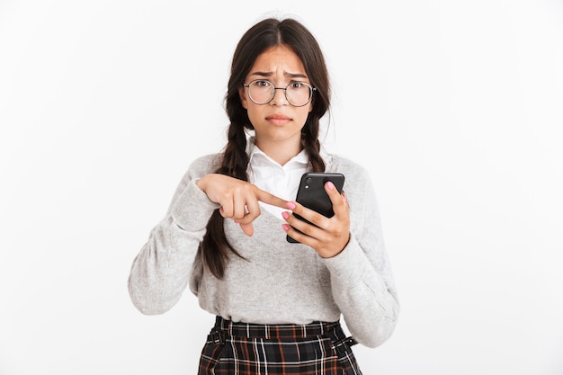 Primer retrato de una adolescente disgustada con anteojos y uniforme escolar expresando perplejidad mientras sostiene el teléfono inteligente aislado sobre la pared blanca