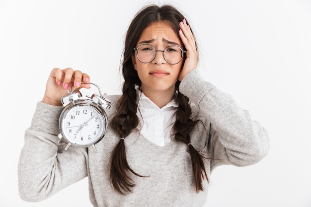 Primer retrato de una adolescente preocupada con anteojos frunciendo el ceño mientras sostiene el despertador en las manos aisladas sobre la pared blanca