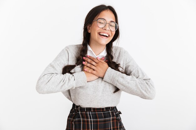 Primer retrato de adorable adolescente con anteojos y uniforme escolar riendo con tocar su pecho aislado sobre la pared blanca