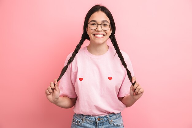 Foto primer retrato de amigable niña feliz con dos coletas con anteojos divirtiéndose y sonriendo al frente aislado sobre pared rosa