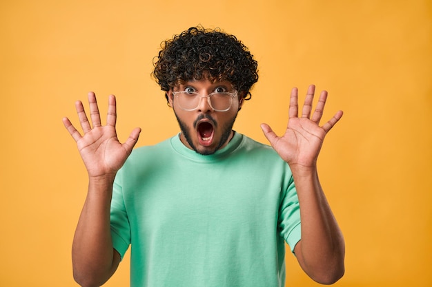 Primer retrato de un apuesto hombre indio con camiseta turquesa y gafas con ojos grandes y boca abierta parado sobre fondo amarillo y expresando emoción de sorpresa y mostrando las manos