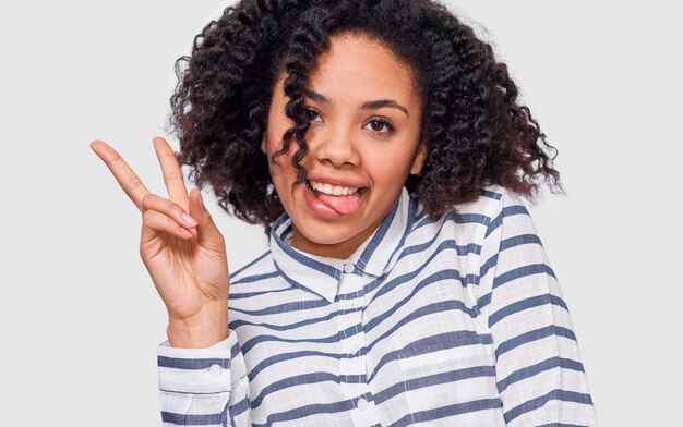 Primer retrato de estudio de una joven afroamericana positiva que sonríe ampliamente mostrando un gesto de paz y muestra la lengua mientras mira a la cámara de pie contra el fondo blanco del estudio